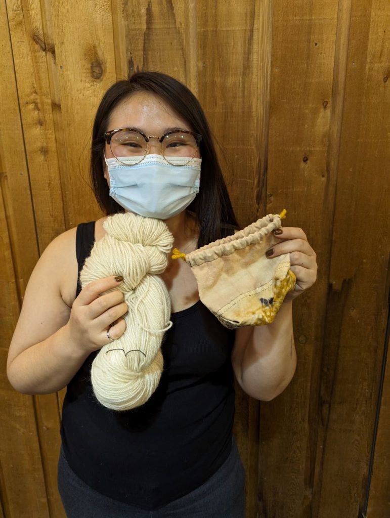 Woman wearing glasses and a mask is proudly holding up a skein of cream colored yarn she spun and a small drawstring bag she sewed with thread she spun.